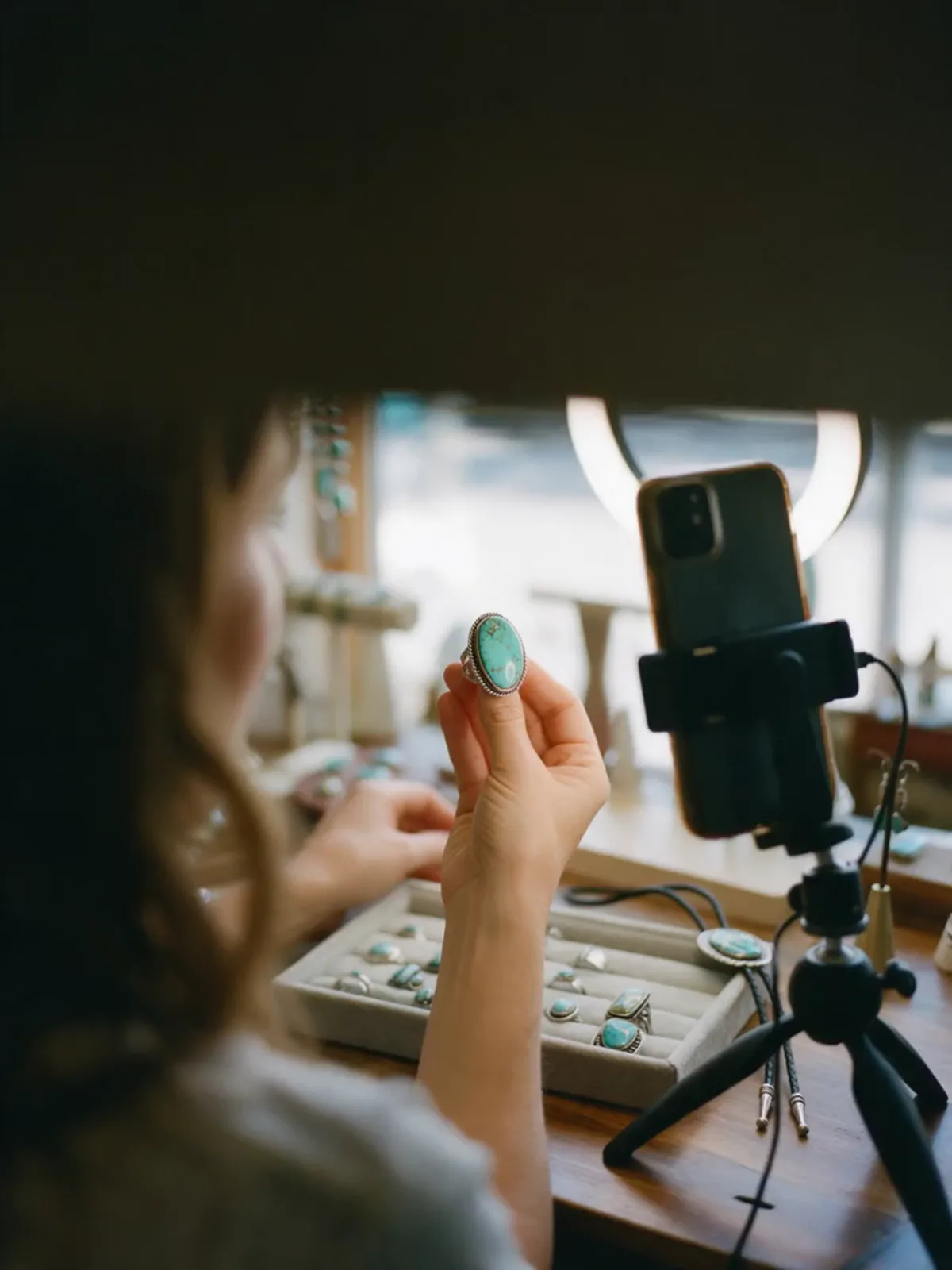 A live-selling host holds a turquoise ring up to her phone on a tripod, seen from behind her shoulder.